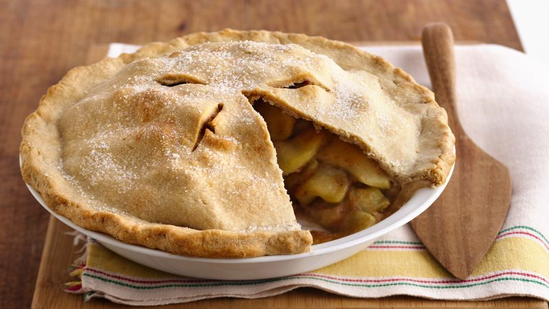 This is a picture on the finished apple pie. There is a slice in the front missing, and you can see the apples showing. The apple pie is sitting on top of a striped place setting, and there is a wood pie slicer sitting next to it.