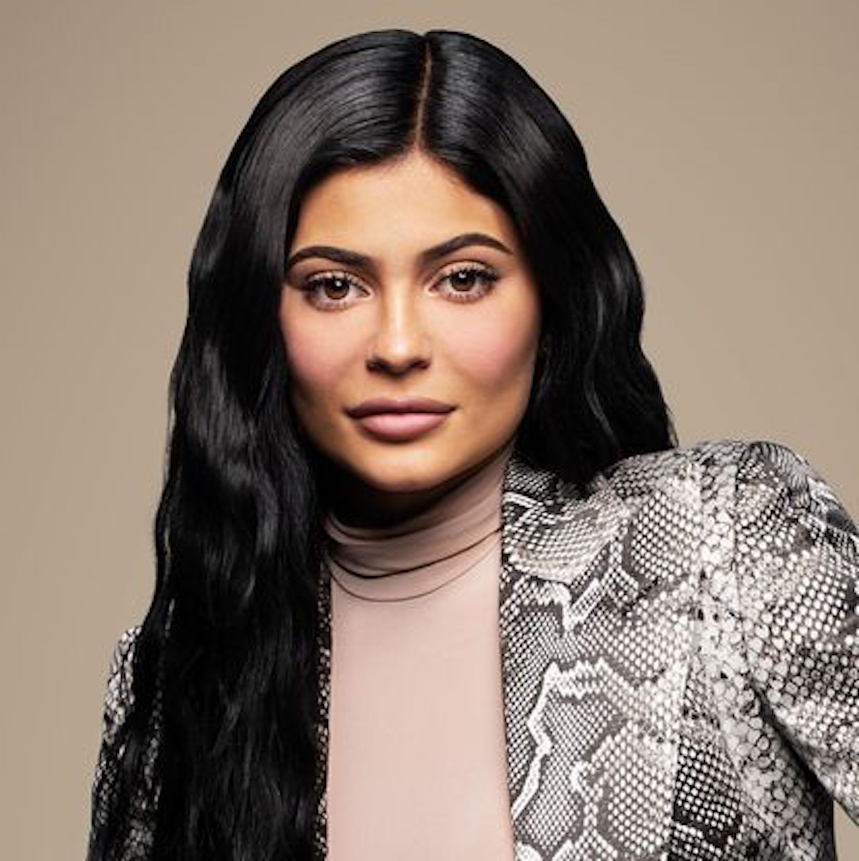 A headshot of Kylie Jenner. She is wearing a light pink turtleneck shirt with a snakeskin blazer over. Kylie is staring directly at the camera, with her long black hair over one shoulder.